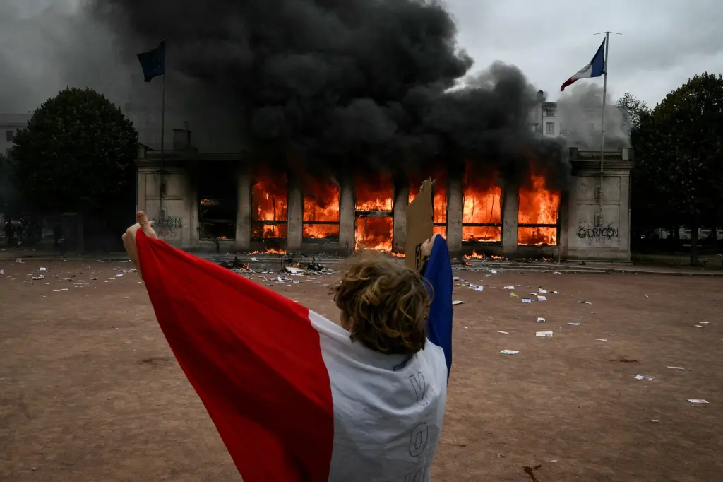 A protester holds a French flag in front of a burning building during riots in France
