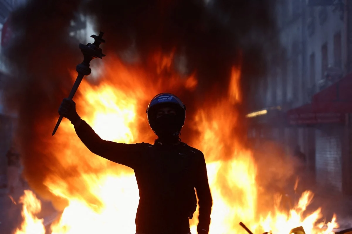 A masked demonstrator holds up an object in front of a burning street barricade during clashes in France